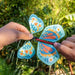 Colorful butterfly-shaped wind spinner held by hands with a green outdoor background