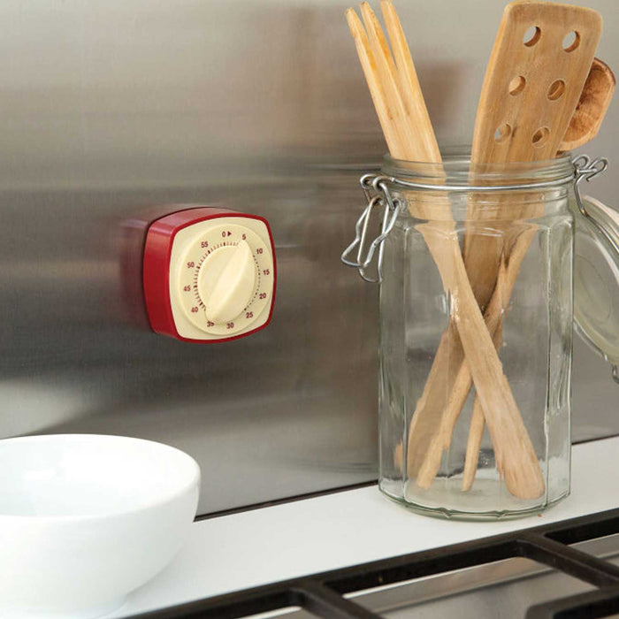 magnetic retro kitchen timer with large cream dial on front and red numbers, and red framed edge. Timer is on a metal kitchen wall with wooden utensils in glass jar to right