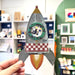 A hand holds a decorative metal rocket-shaped wall hanging with a textured galvanized finish, a circular green window, a red-and-white checkered section, and a yellow-orange flame detail, set against a blurred indoor shop background.