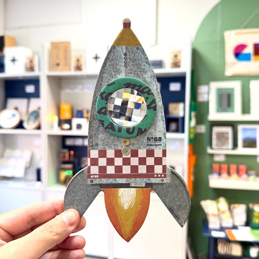 A hand holds a decorative metal rocket-shaped wall hanging with a textured galvanized finish, a circular green window, a red-and-white checkered section, and a yellow-orange flame detail, set against a blurred indoor shop background.