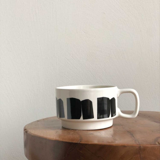 white ceramic cup with 5 black rectangular shapes around the outside. Cup is on a wooden table top with grey wall in background