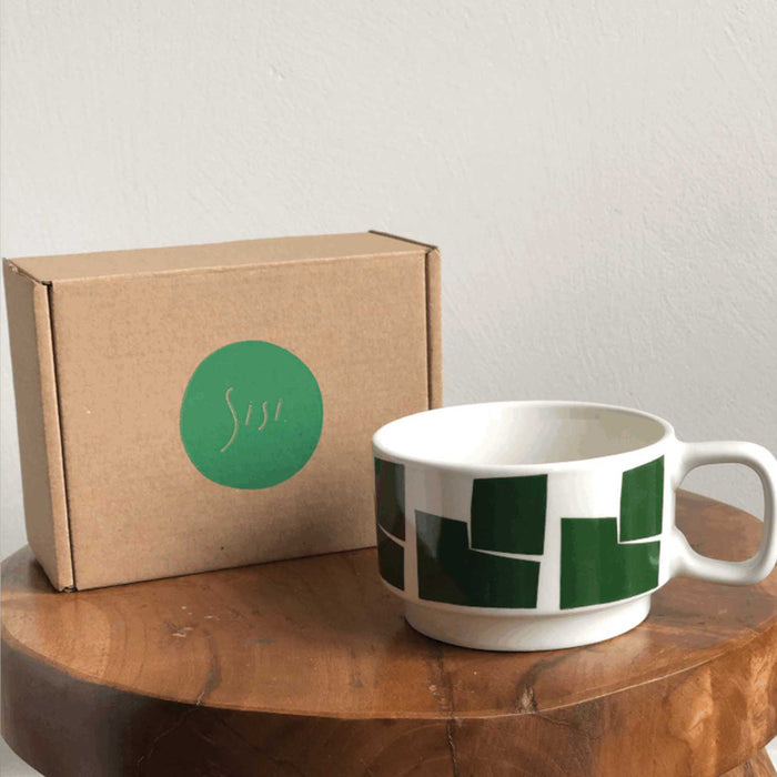 white ceramic cup with 5green abstract shapes around the outside. Cup is on a wooden table top with a cardboard box with green circle on it behind cup, grey wall in background