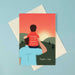 greeting card with young boy sitting on a mans right shoulder with his arm on the man's head. they have their back to us and are watching a sunset over green hills with a red sky 