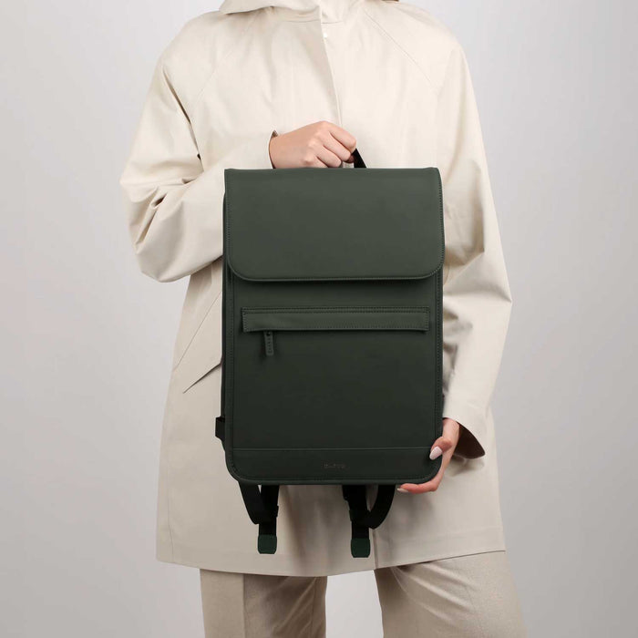 woman in beige raincoat and trousers holding a green backpack in front of her
 