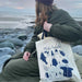 woman sitting on rocks on beach looking to sea with green coat and hat on holding a white and blue tote on her shoulder
