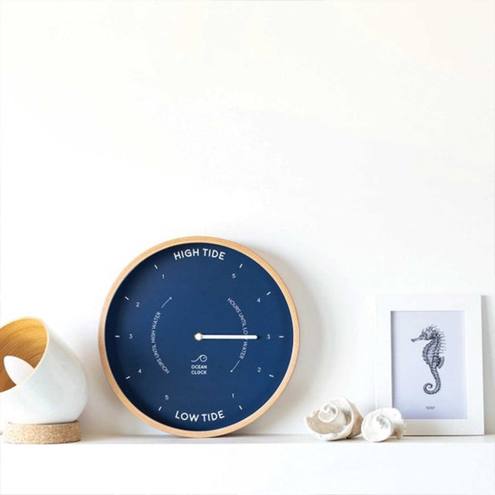 navy round tide clock with wooden frame on a white shelf with between a white lamp and framed picture of a seahorse