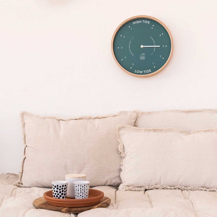 A teal wall clock with a tide indicator, encased in a natural beech wood frame, hanging on a white wall above a beige sofa with cushions. The clock shows markings for high and low tide.