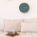 A teal wall clock with a tide indicator, encased in a natural beech wood frame, hanging on a white wall above a beige sofa with cushions. The clock shows markings for high and low tide.