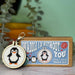 Cross-stitched penguin keychain and 'Waddle I Do Without You' cross-stitch mini kit on a wooden surface with a blurred plant background.