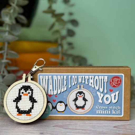 Cross-stitched penguin keychain and 'Waddle I Do Without You' cross-stitch mini kit on a wooden surface with a blurred plant background.