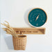 wooden wall shelf with 4 hanging pegs, wicker basket with wheat hanging from left peg and circular wooden weather barometer with a green face sitting on top right of shelf