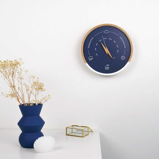 blue faced wall clock with white markiings and dial, wooden and white frame and blue vase to bottom left corner