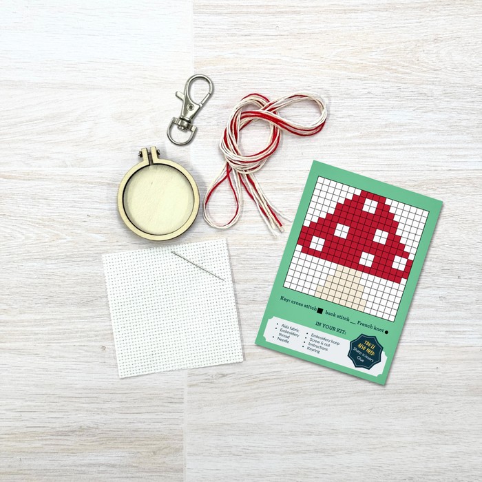 Cross-stitch kit with red space Invader design, hoop, thread, and pattern chart on a light wooden surface.