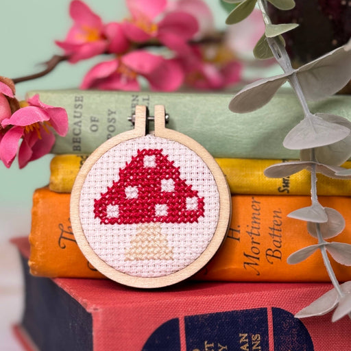 Cross-stitched mushroom design in a wooden hoop on books with flowers and leaves.