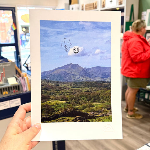 A hand holds a white-bordered art print featuring a mountain landscape under a blue sky with a small smiling cloud and clover graphic. The background shows a real store interior with a person in a red jacket blurred on the right.