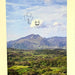 A square art print or card featuring a landscape photograph of green hills and mountains under a blue sky with clouds, with a simple line drawing of a smiling cloud holding a shamrock near the top center.