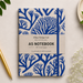 A5 notebook with a blue coral pattern on a cream cover, styled in a flat-lay with a gold pen, baby's breath, and green leaves on a light beige background.
