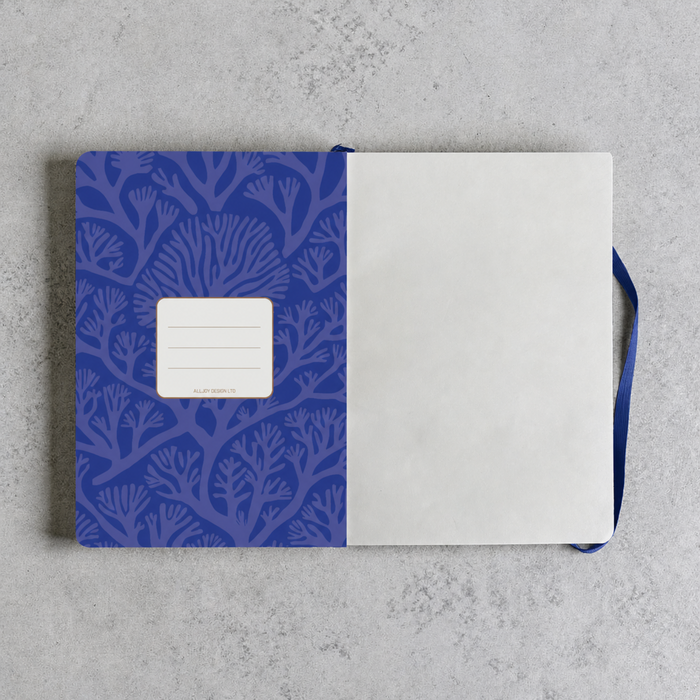 A notebook with a blue patterned cover and blank white pages, laid flat on a light gray textured surface. A blue ribbon bookmark is visible on the right side.