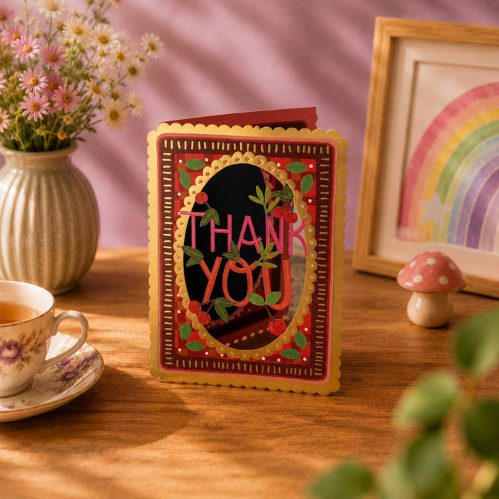 Decorative 'Thank You' card on a wooden table with a cup of tea, flowers, and a rainbow picture frame.