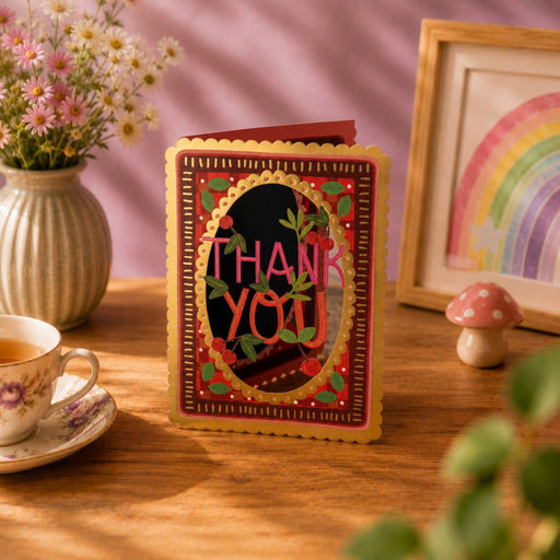 Decorative 'Thank You' card on a wooden table with a cup of tea, flowers, and a rainbow picture frame.