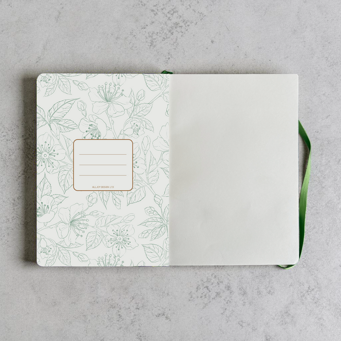 An open notebook or journal laid flat on a light gray textured surface. The left page has a white background with a delicate green botanical line pattern and a small rectangular label area outlined in green.