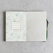 An open notebook or journal laid flat on a light gray textured surface. The left page has a white background with a delicate green botanical line pattern and a small rectangular label area outlined in green.