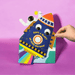 Animation showing a hand adjusting the jiggly eyes on a colourful paper card of a rocket ship and an astronaut on a planet with a pink background. 