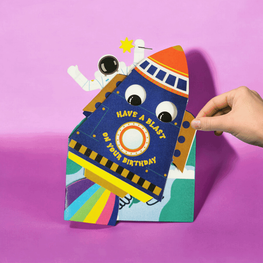 Animation showing a hand adjusting the jiggly eyes on a colourful paper card of a rocket ship and an astronaut on a planet with a pink background. 