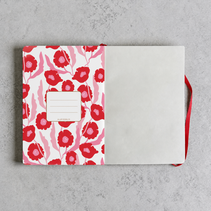 An open notebook with a white cover decorated with a red and pink floral pattern, featuring a blank name label on the front and a red ribbon bookmark, set against a textured gray background.
