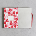An open notebook with a white cover decorated with a red and pink floral pattern, featuring a blank name label on the front and a red ribbon bookmark, set against a textured gray background.