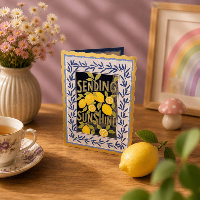 Greeting card with lemons and 'sending sunshine' text on a table with a lemon, teacup, and flowers.