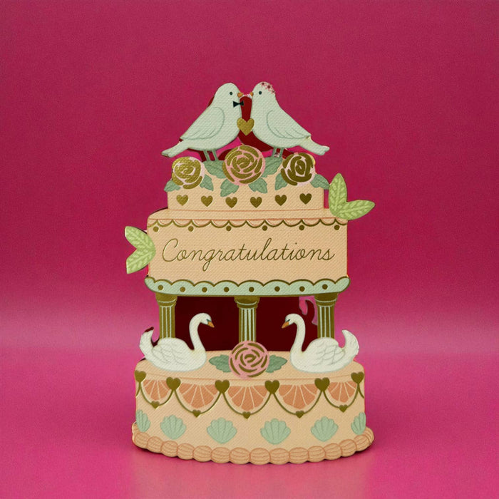 A card in the shape of a decorative tiered cake with two white birds perched on top, flowers on the upper tier, the word 'Congratulations' across the center, and two swans on the lower tier, set against a pink background.