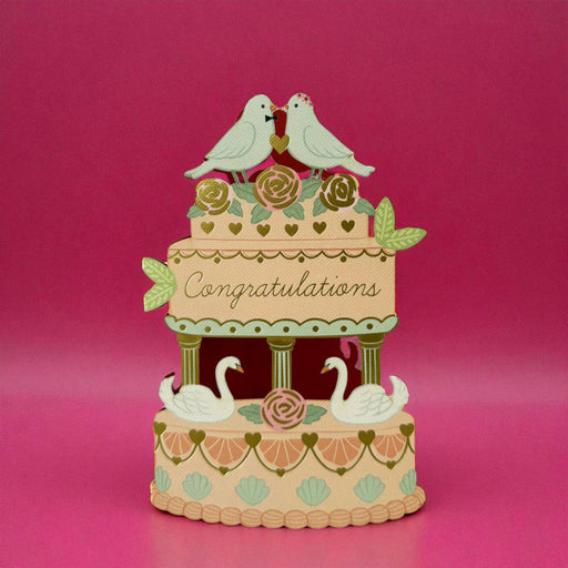 A card in the shape of a decorative tiered cake with two white birds perched on top, flowers on the upper tier, the word 'Congratulations' across the center, and two swans on the lower tier, set against a pink background.