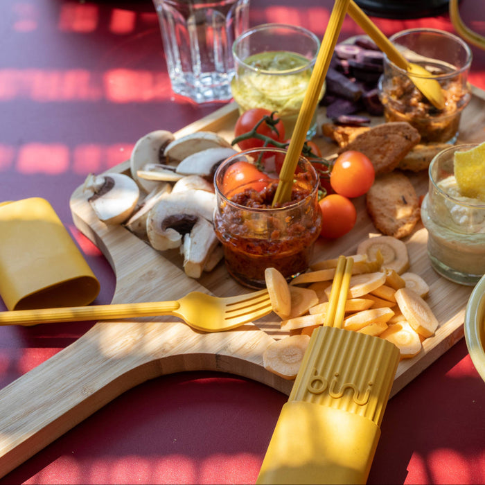 Assorted snacks on a wooden board with a bowl of chips on a red tablecloth.