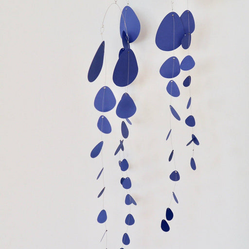 A minimalist hanging mobile made of layered, irregular blue teardrop and oval shapes suspended from thin metal wires against a plain off-white wall.