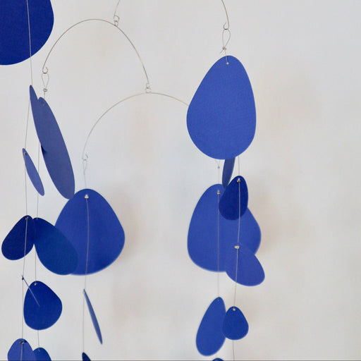 A minimalist metal wire hanging mobile with irregular blue paper cutouts, suspended against a plain off-white wall.