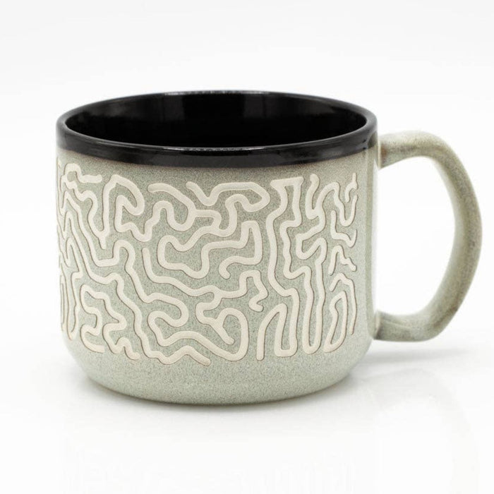A ceramic mug with a light sage green exterior featuring a raised cream maze pattern, a solid black interior, and a matching sage green handle, shown on a white background.