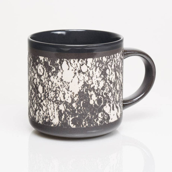 A ceramic mug with a glossy black finish and a textured, splattered white pattern on the exterior, shown on a reflective white surface against a white background.