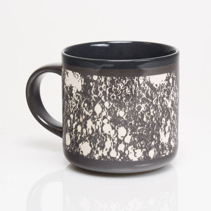 A ceramic mug with a glossy black interior and handle, featuring a textured black and white speckled pattern on the exterior, shown on a reflective white surface.