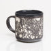A ceramic mug with a glossy black interior and handle, featuring a textured black and white speckled pattern on the exterior, shown on a reflective white surface.
