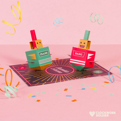 A pop-up cardboard party decoration set featuring two box-like characters labeled CRASH and BANG standing on a circular black and pink base, with colorful confetti and streamers scattered on a pink background. The Clockwork Soldier logo appears in the bottom right corner.