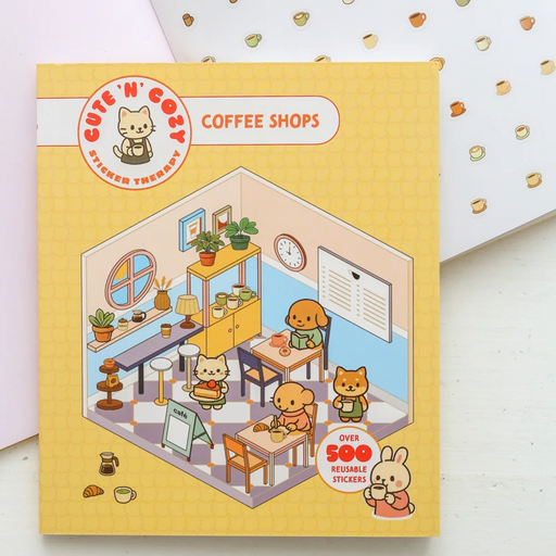 A yellow 'Cute 'n Cozy Coffee Shops' sticker therapy kit box shown on a white surface with a pink envelope behind it. The box features a detailed isometric illustration of a cozy animal cafe interior with cute animal characters, tables, chairs, plants, and shelves.