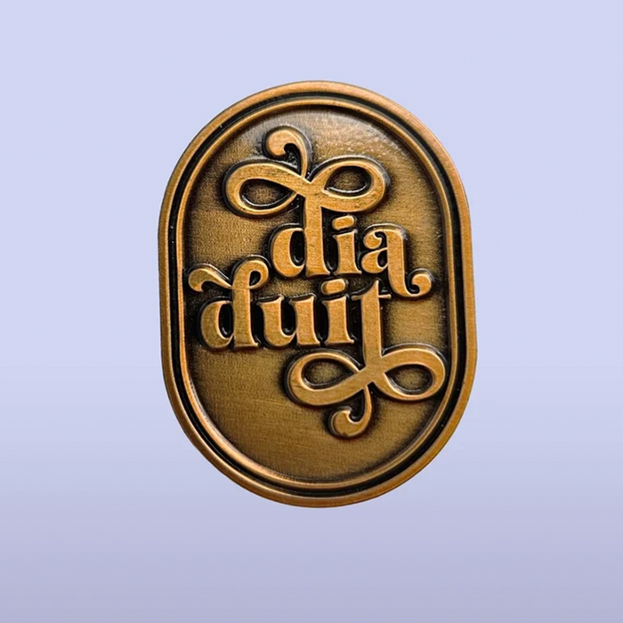 An oval bronze-colored metal plaque or emblem with raised gothic-style lettering reading 'dia duit' on a soft lavender gradient background.