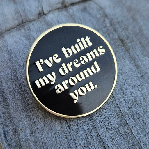 A round black enamel pin with a gold-toned border and raised white text reading 'I've built my dreams around you.' placed on a weathered gray wood surface.