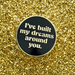 A round black enamel pin with a gold border and white text reading 'I've built my dreams around you.' placed on a sparkling gold glitter background.