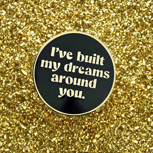 A round black enamel pin with a gold border and white text reading 'I've built my dreams around you.' placed on a sparkling gold glitter background.