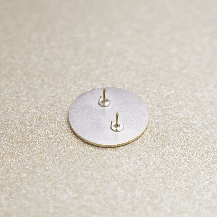 A close-up of the back of a round silver-toned earring, showing two post and butterfly backings, placed on a textured beige surface.