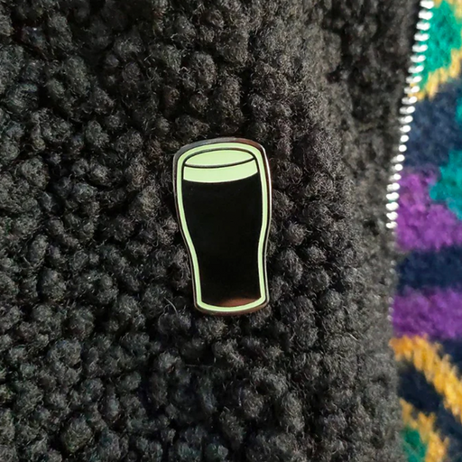 A small enamel pin shaped like a dark pint glass with a glowing pale green outline, placed on a black textured fleece surface next to a colorful knit fabric.