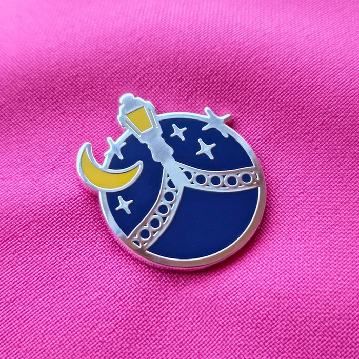 A circular enamel pin featuring a blue night sky with white stars, a yellow crescent moon, and a silver street lamp, displayed on a textured hot pink fabric background.