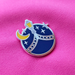 A circular enamel pin featuring a blue night sky with white stars, a yellow crescent moon, and a silver street lamp, displayed on a textured hot pink fabric background.